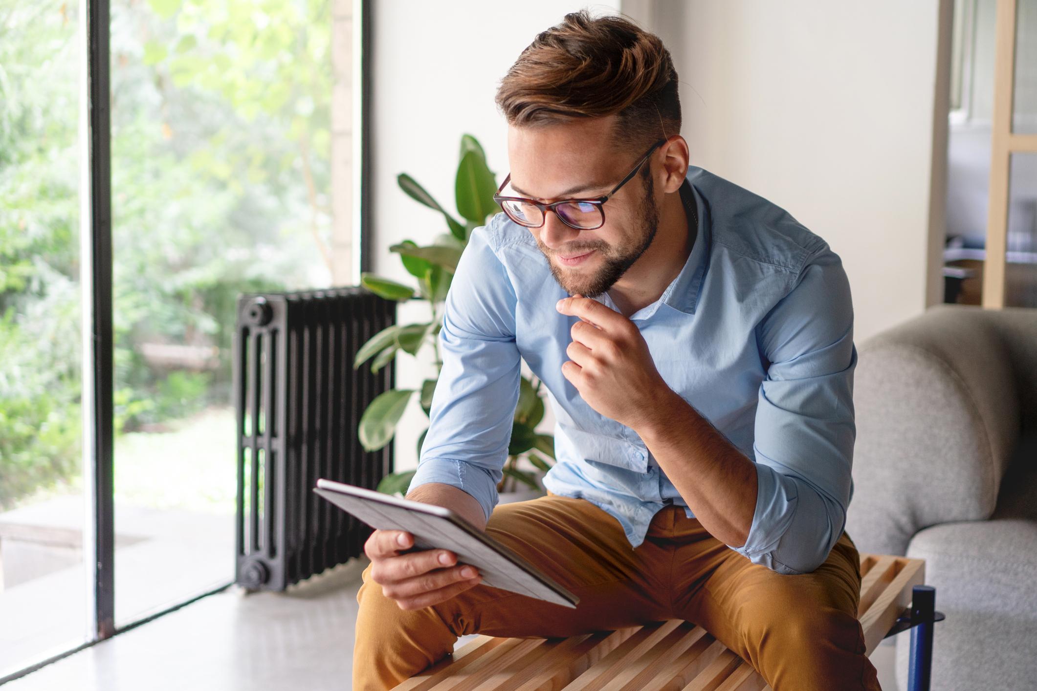 A man is sitting on a bench as he reads from a digital tablet. 